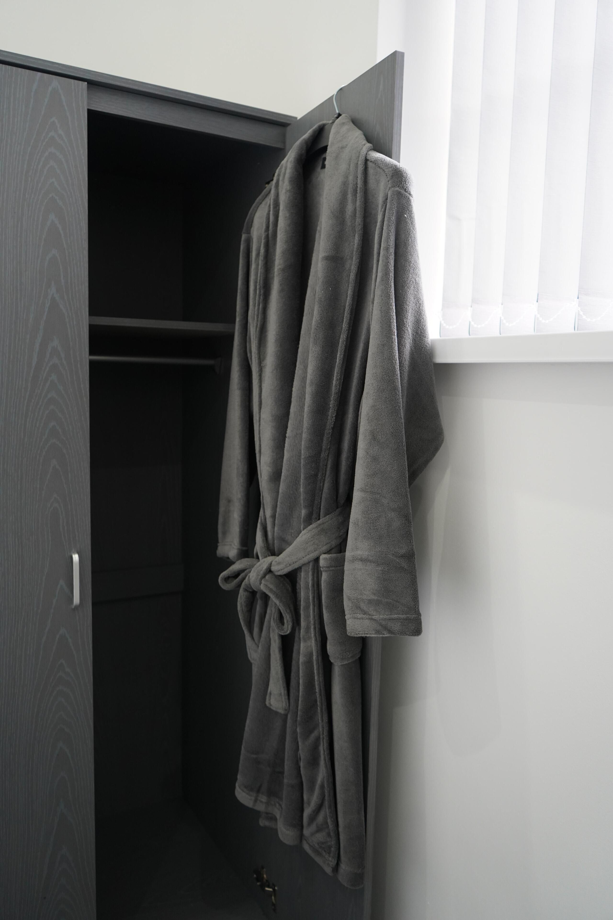 Una bata gris colgada de un perchero en una habitación con un armario y una ventana con persianas.