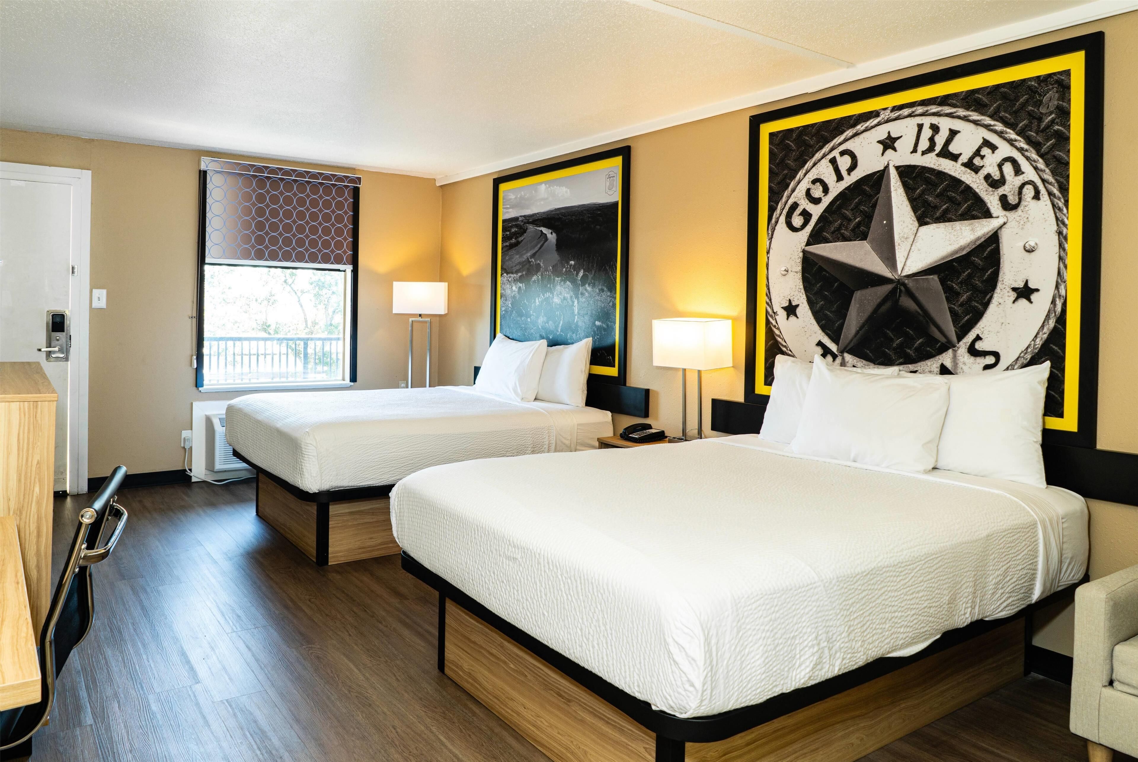 Une chambre d’hôtel avec deux lits, une grande fenêtre et un mur orné d’une inscription « Que Dieu vous bénisse ».