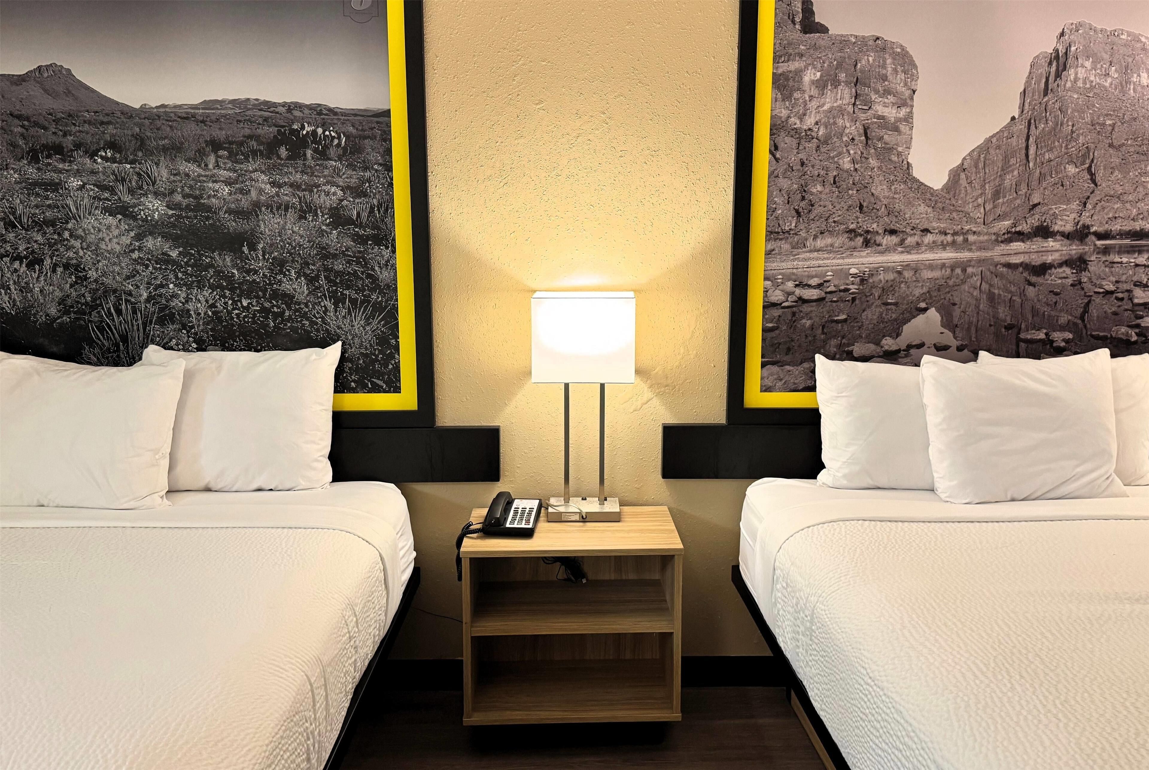 Habitación de hotel con dos camas, una mesita de noche con lámpara, un teléfono y un mural en la pared que representa un paisaje desértico.