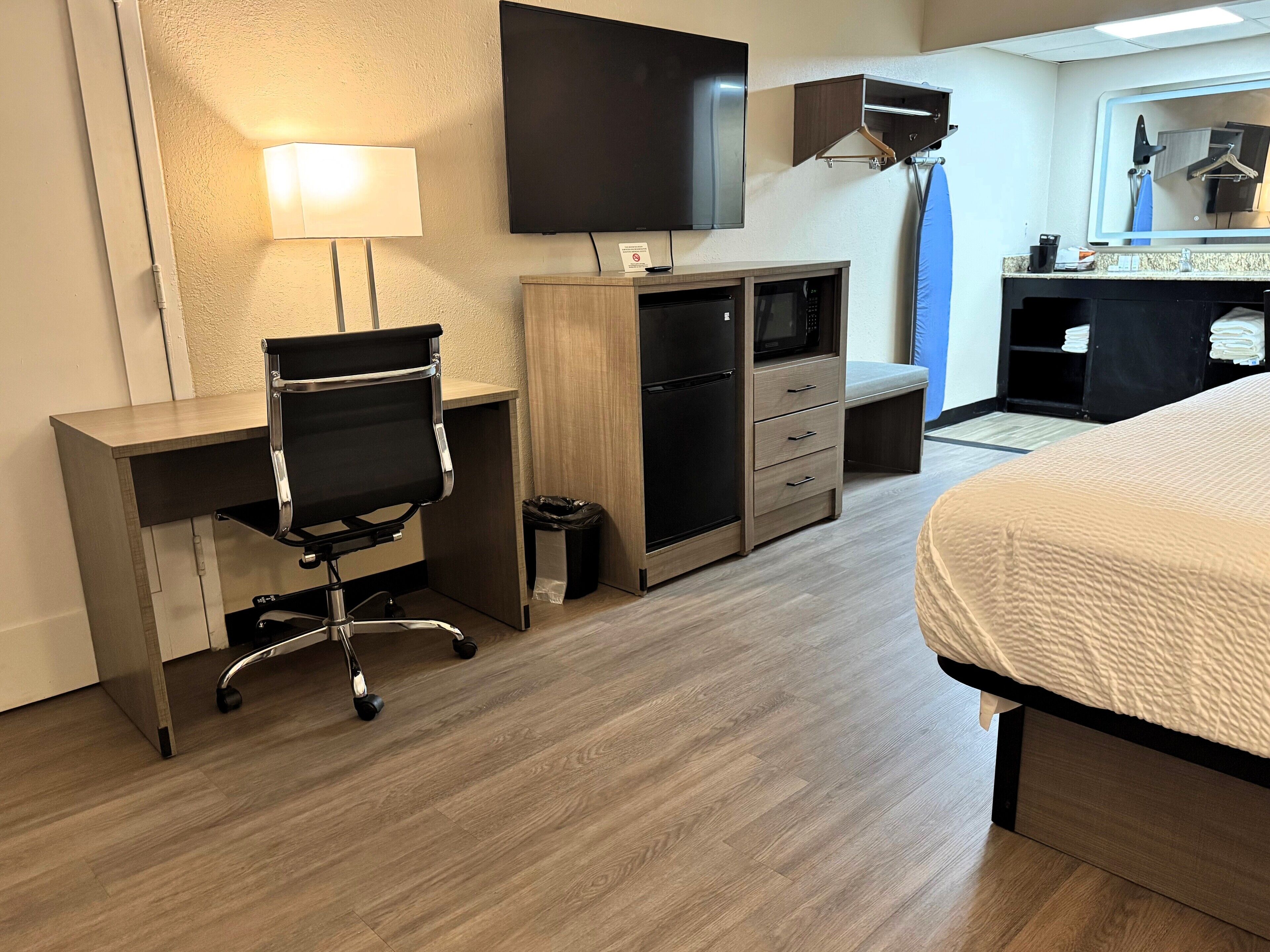 A hotel room with a desk, chair, bed, TV, and a small kitchenette.
