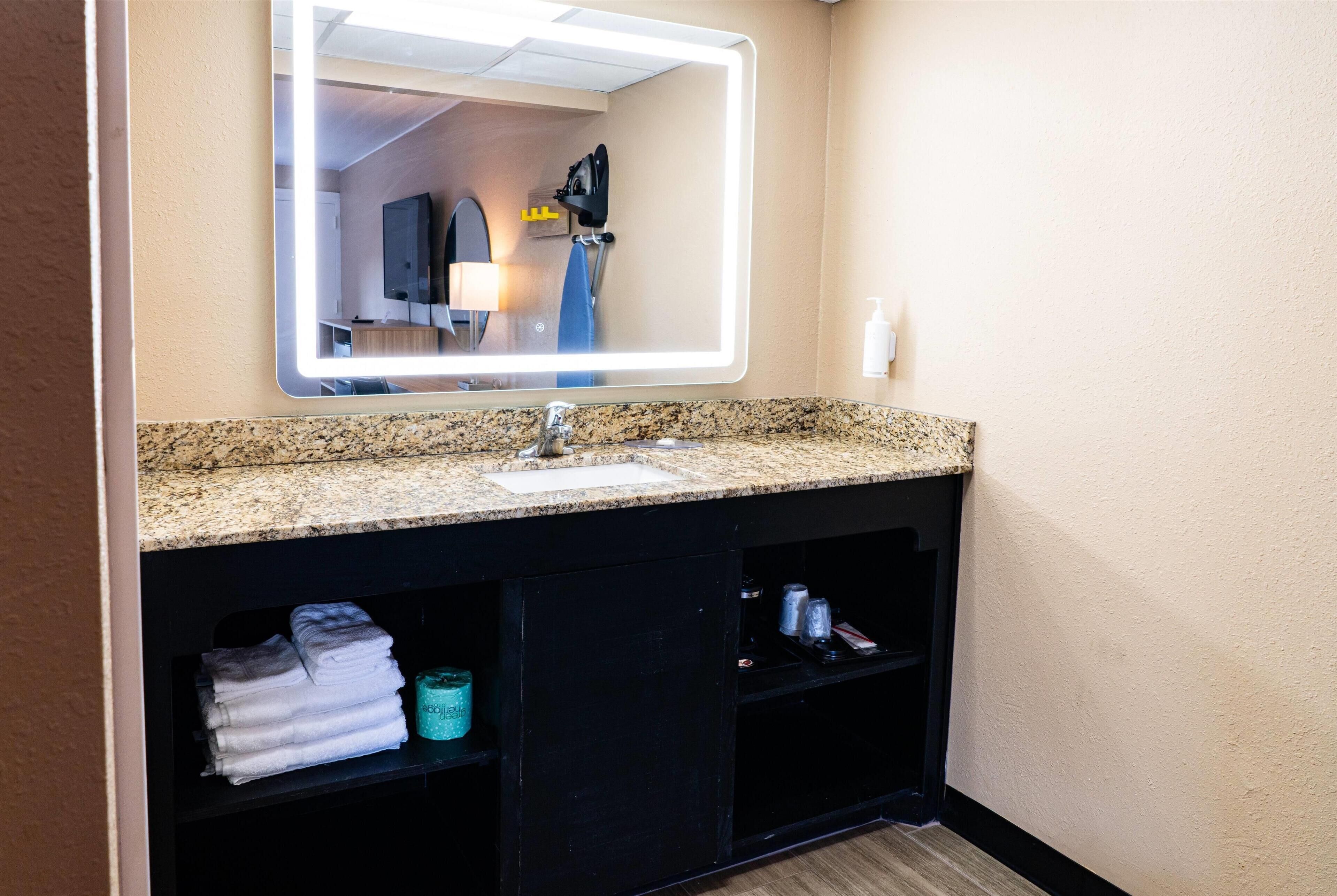 A bathroom with a granite countertop, a sink, a mirror, and a shelf with folded towels and toiletries.