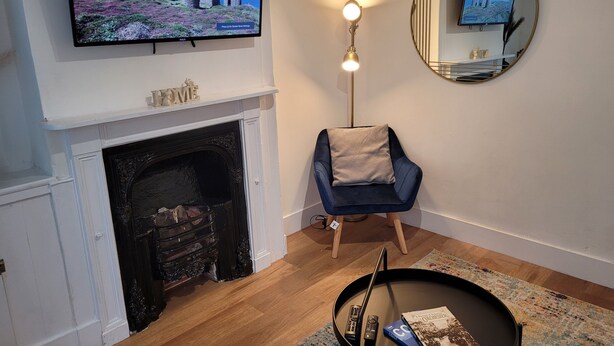 Una sala de estar con chimenea, un televisor montado encima, un espejo redondo en la pared, un sillón azul y una mesita con un libro y controles remotos.