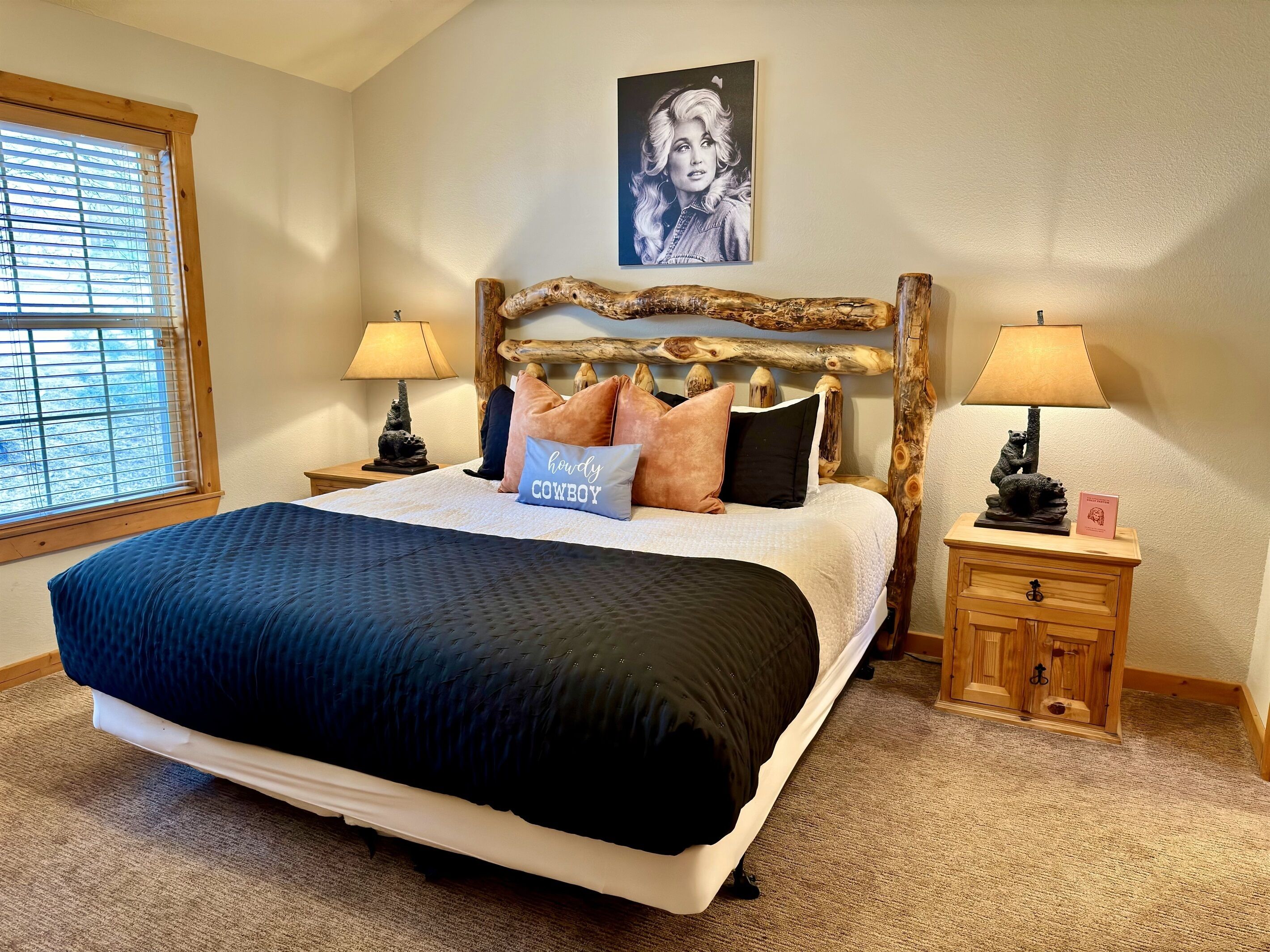 Een slaapkamer met een groot bed, twee nachtkastjes, een houten hoofdbord, een raam met jaloezieën en een aan de muur hangend schilderij.