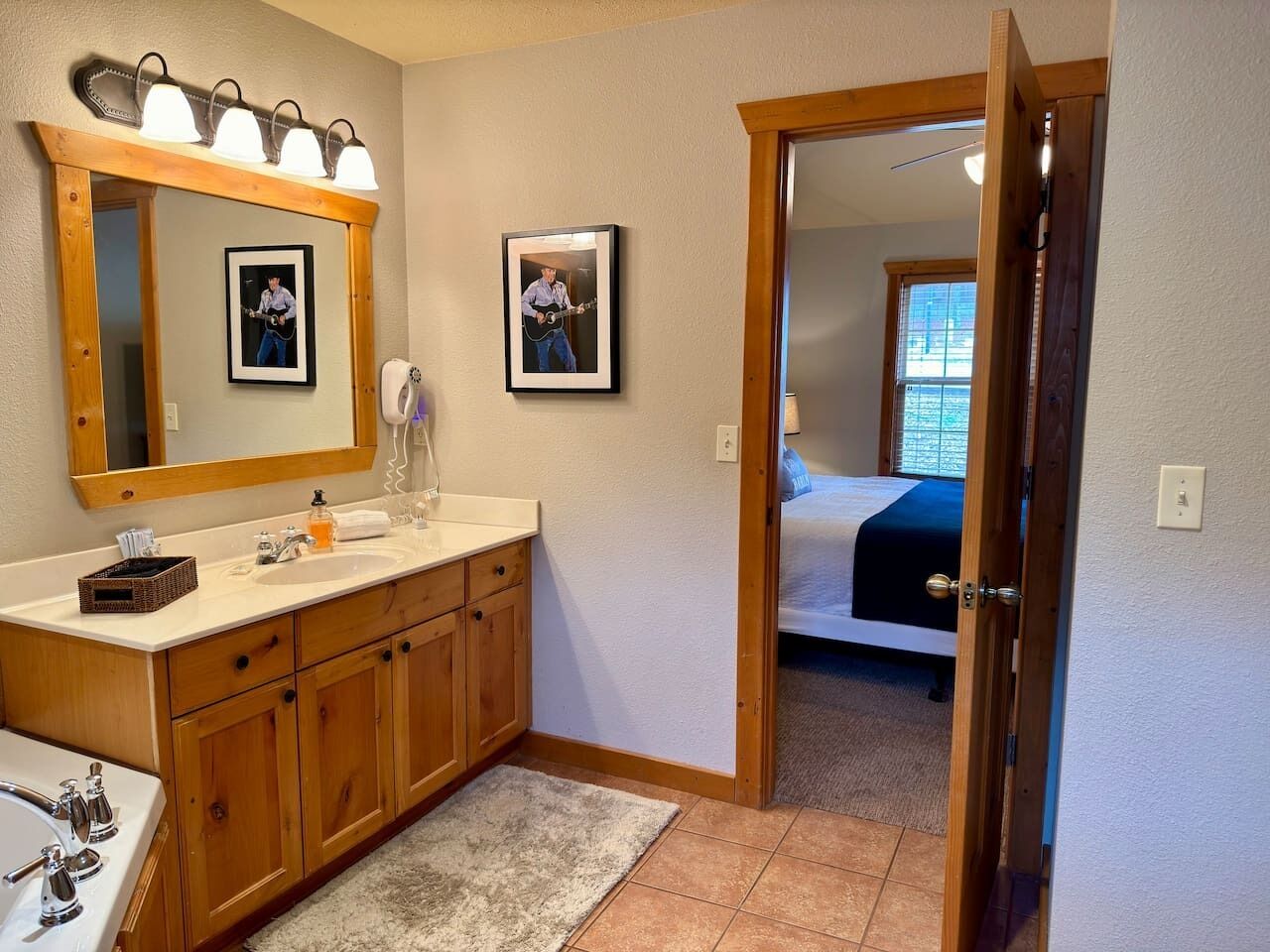 A bathroom with wooden cabinets, a sink, and a mirror. There are framed pictures on the wall and a door leading to a bedroom.