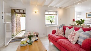 A living room with a red sofa, a wooden coffee table, and a vase with flowers. There is a window with a view of greenery outside. - Lavender Cottage -  a cottage that sleeps 2 guests  in 1 bedroom (Portscatho)