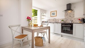 A modern kitchen with white cabinetry, a built-in oven, and a dining area with a wooden table and white chairs. - Lavender Cottage -  a cottage that sleeps 2 guests  in 1 bedroom (Portscatho)