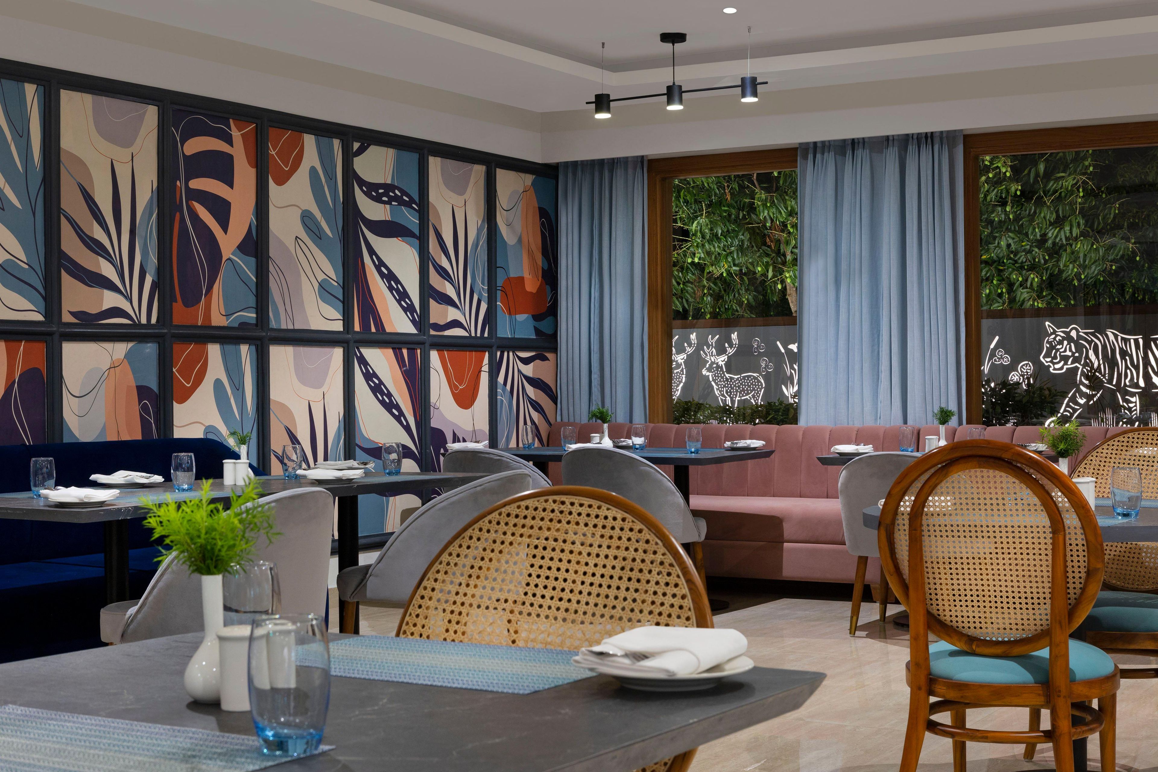 A modern restaurant with a vibrant abstract mural, comfortable seating, and a view of greenery outside.
