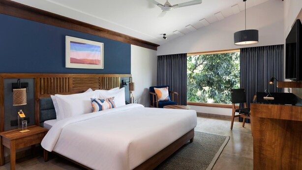 A hotel room with a large bed, a desk, a chair, a ceiling fan, and a window with curtains. - Voco Jim Corbett (Ramnagar)