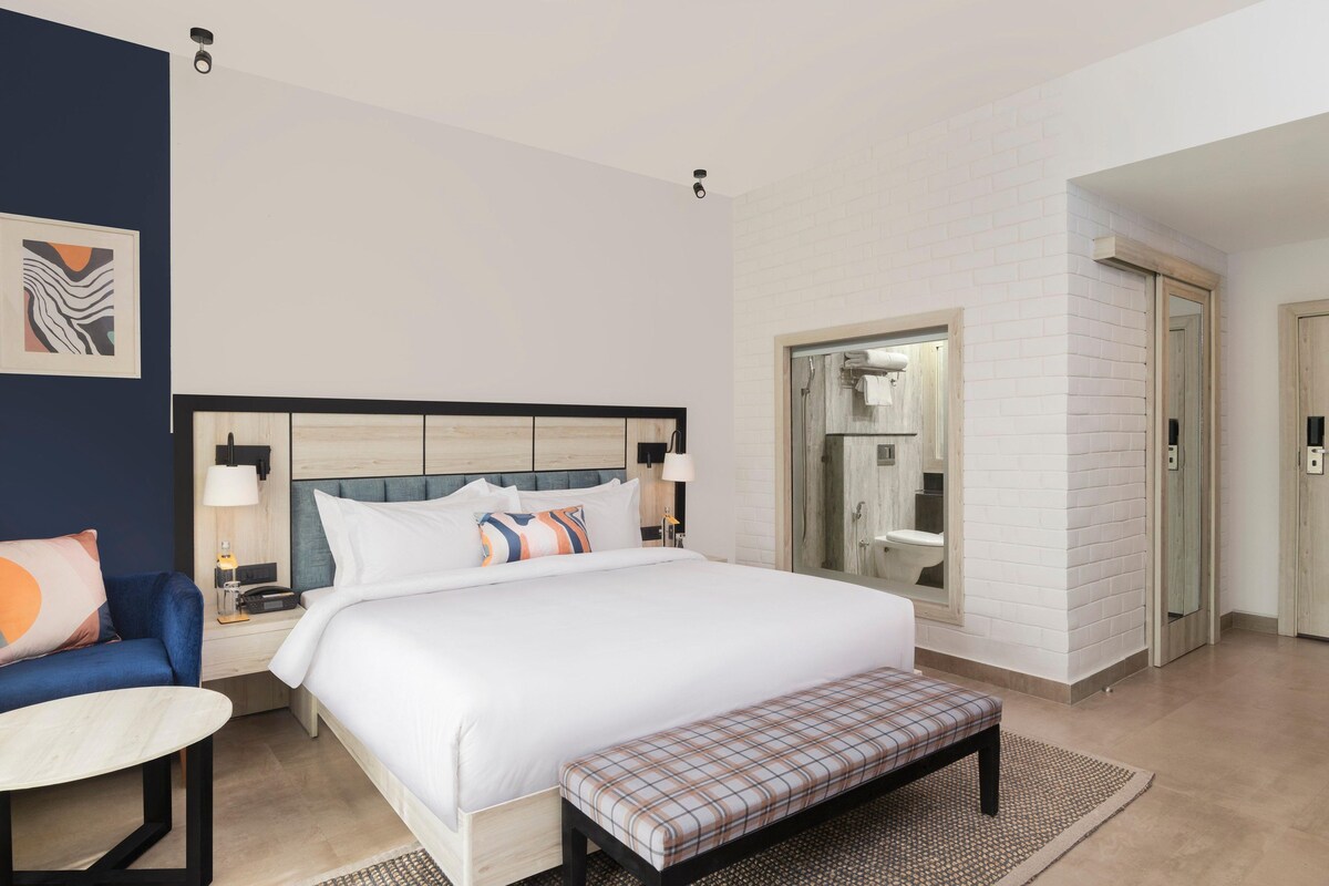 A hotel room with a large bed, a bedside table, a chair, a bench, a mirror, and a bathroom with a shower and toilet.