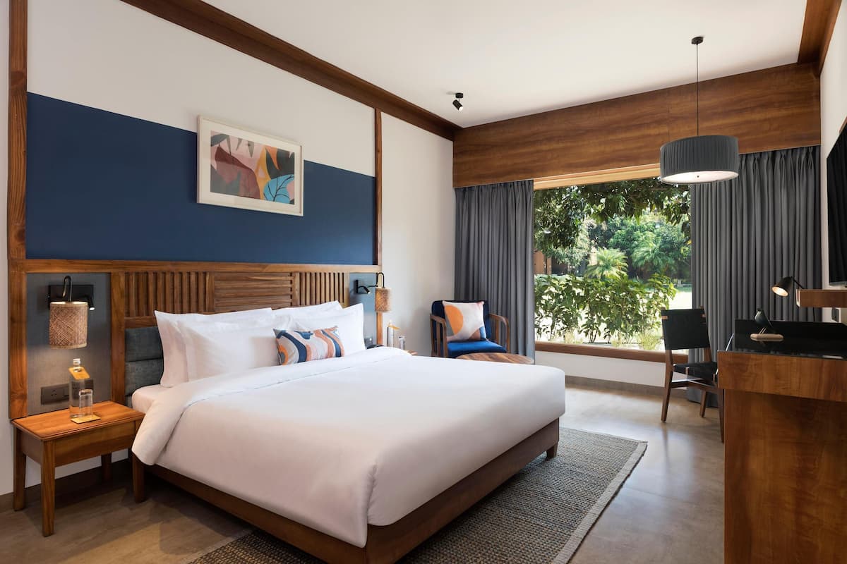 A modern hotel room with a large bed, wooden headboard, a desk, and a view of greenery through the window.