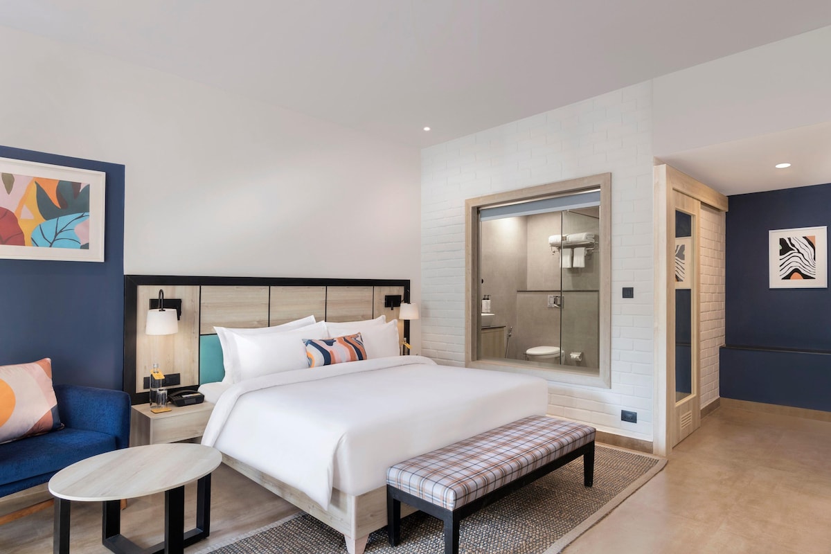 A modern hotel room with a large bed, bedside tables, a bench, a small table, and a bathroom with a glass shower enclosure.