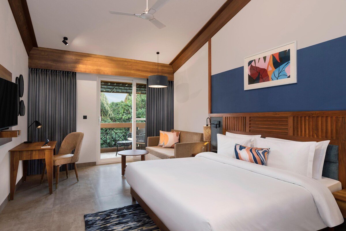 Une chambre d’hôtel avec un lit, un bureau, une chaise, un canapé et une télévision. Il y a une fenêtre avec des rideaux et une vue sur la verdure à l’extérieur.