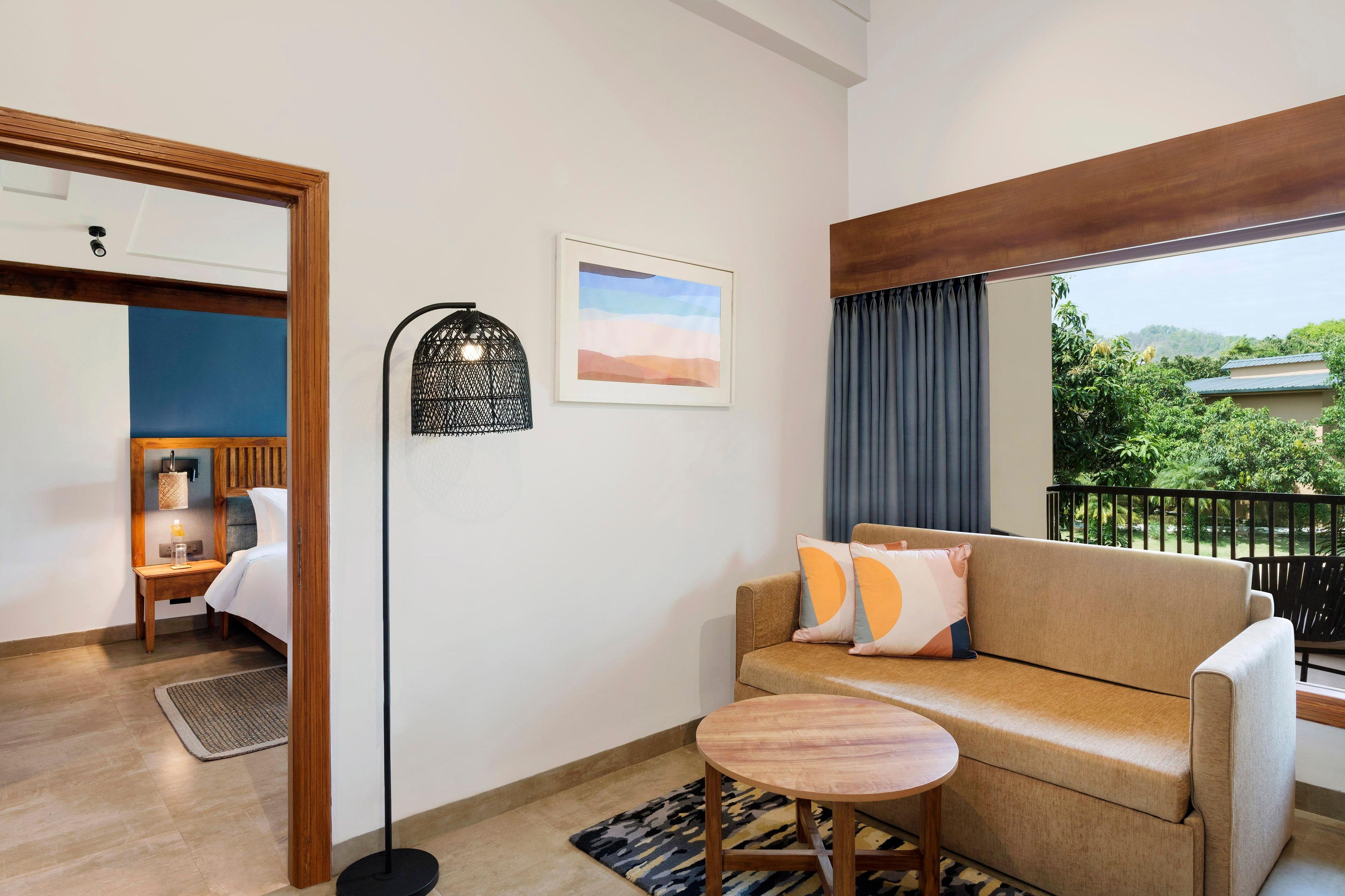 A hotel room with a sofa, a small round table, a floor lamp, a bed, a nightstand, a picture on the wall, and a balcony with a view of greenery.