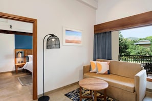 Hotelkamer met een bank, een klein rond tafeltje, een vloerlamp, een bed, een nachtkastje, een foto aan de muur en een balkon met uitzicht op groen.