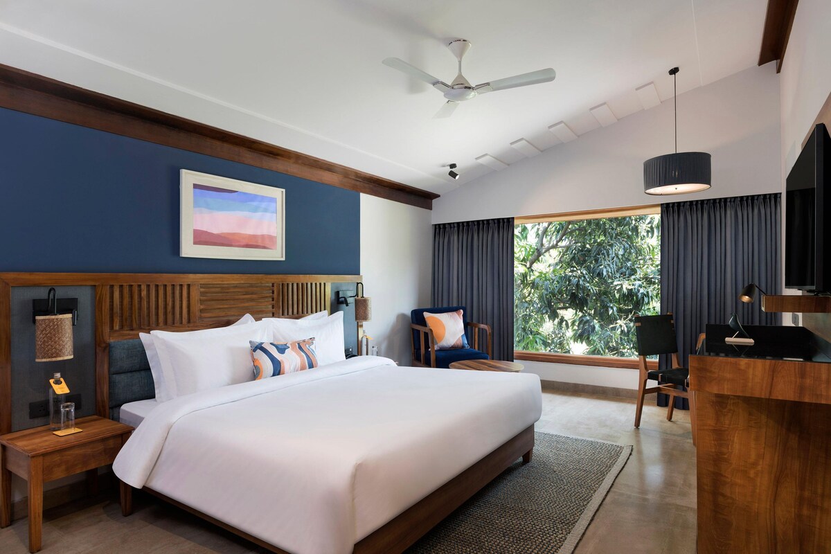 Une chambre d’h ôtel avec un grand lit, un bureau, une chaise, un ventilateur de plafond et une fenêtre avec des rideaux.