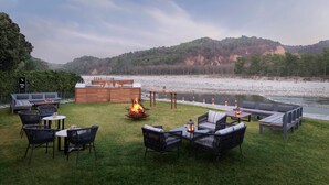 An outdoor seating area with a fire pit, tables, and chairs, overlooking a river and hills. - Voco Jim Corbett (Ramnagar)