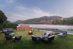 An outdoor seating area with a fire pit, tables, and chairs, overlooking a river and hills.