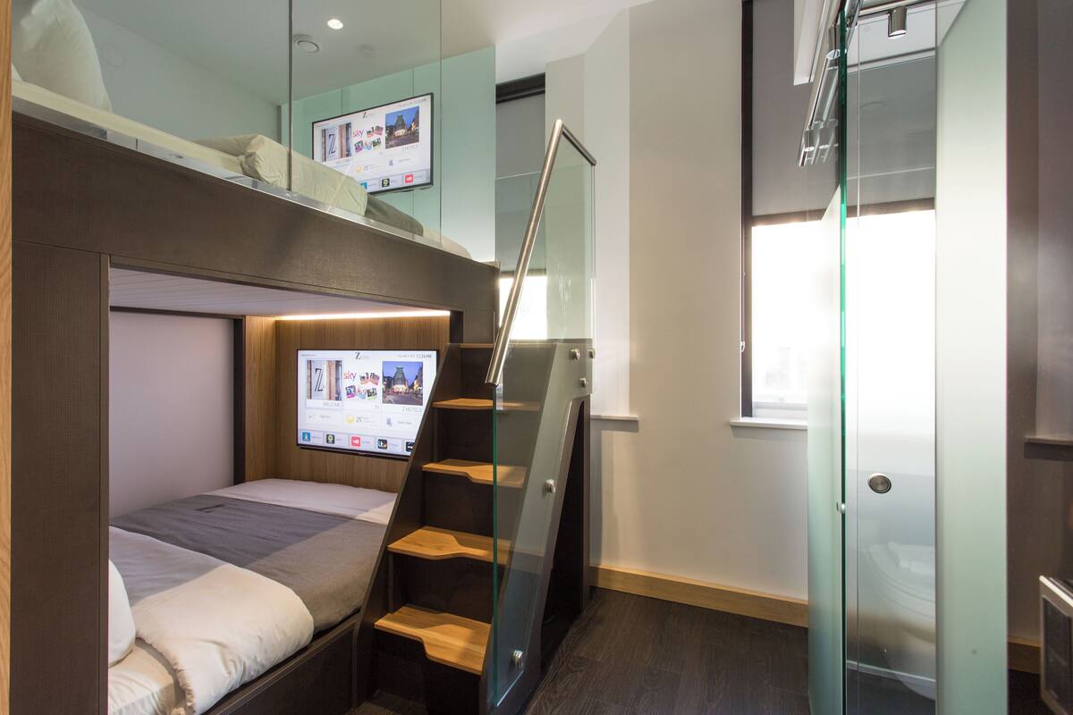 A compact hotel room with a bunk bed, a glass staircase, and a digital screen displaying travel information.