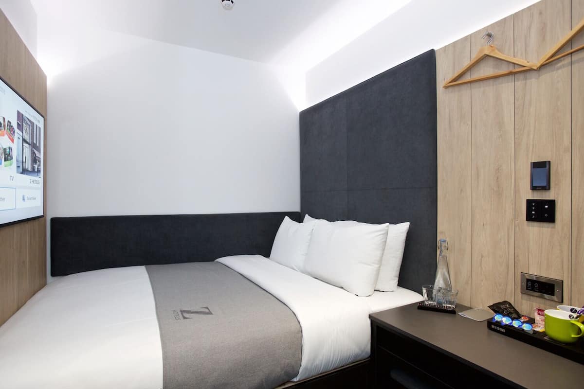 A modern hotel room with a large bed, a TV mounted on the wall, a wooden panel wall, a bedside table with a remote control, a glass, and a cup.