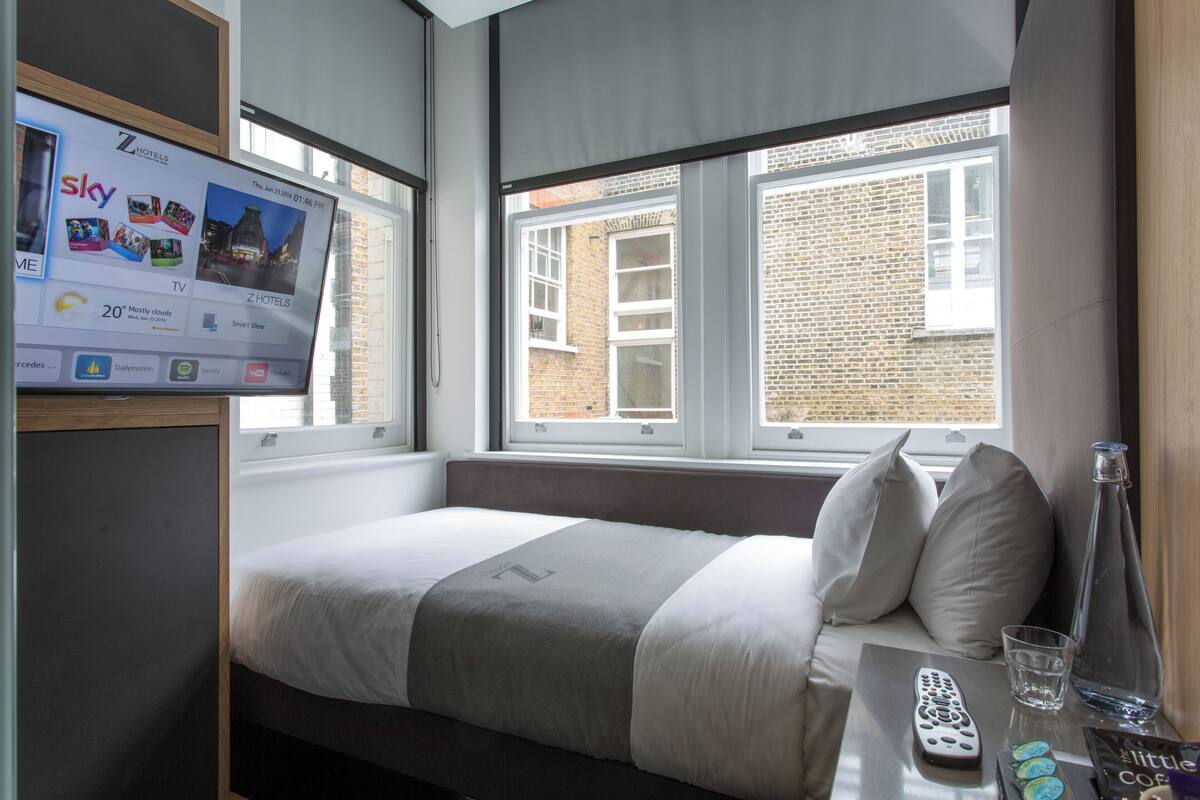 A hotel room with a bed, a TV displaying a hotel channel, a window with a view of a brick building, and a remote control on a table.
