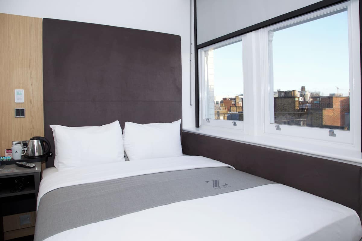 A hotel room with a large bed, a wooden headboard, a window with a city view, and a desk with a kettle and coffee cups.