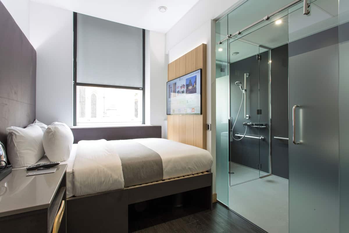 A modern hotel room with a bed, a desk with a remote control, a large window, a glass shower enclosure, and a mounted TV displaying a travel website.
