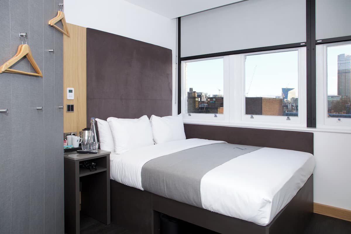 A modern hotel room with a large bed, a bedside table, a wardrobe, and a window with a city view.