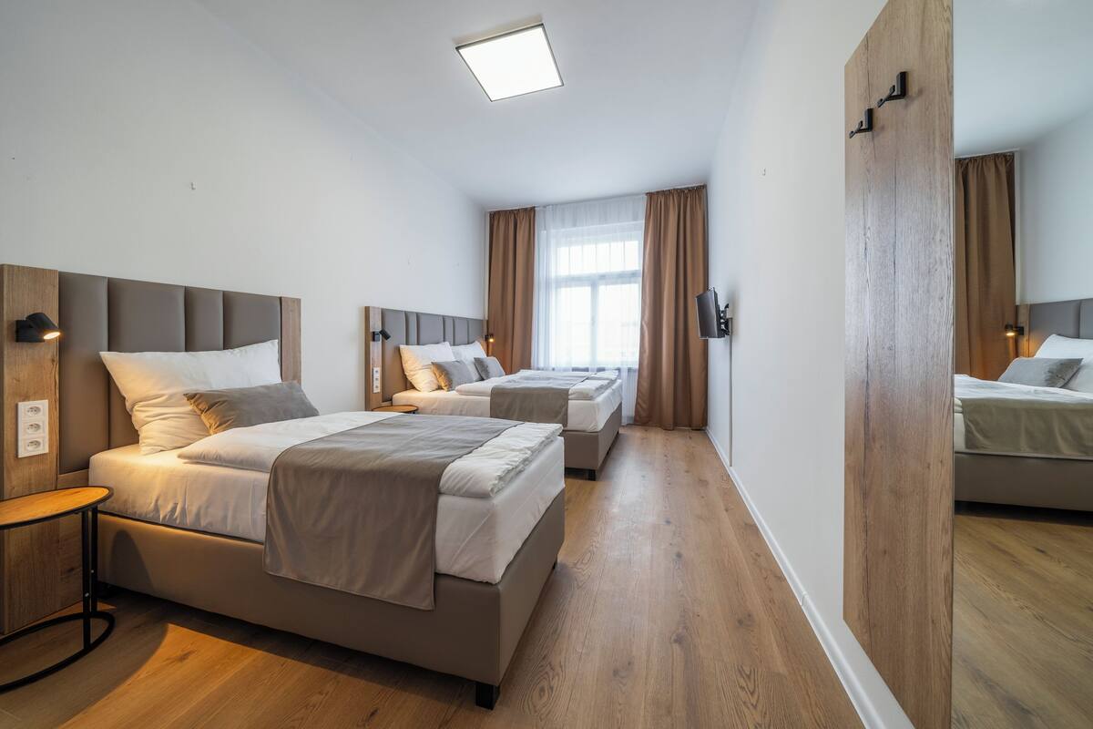 A modern hotel room with a large bed, a smaller bed, a desk, and a window with curtains.