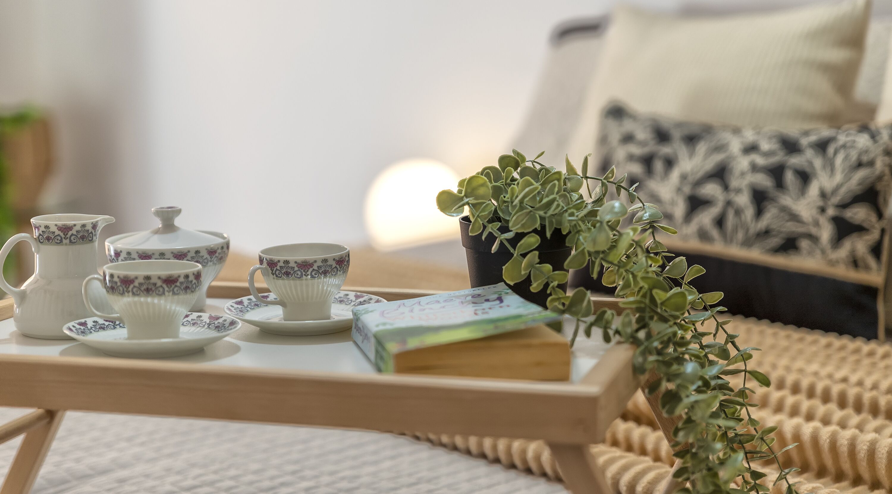 Uma bandeja com jogo de chá e um livro sobre uma mesa em um quarto aconchegante, com um sofá estampado e uma planta.