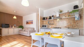 A modern kitchen with white cabinets, a microwave, and a sink. There is a dining area with a table set for two, and a sofa in the background.