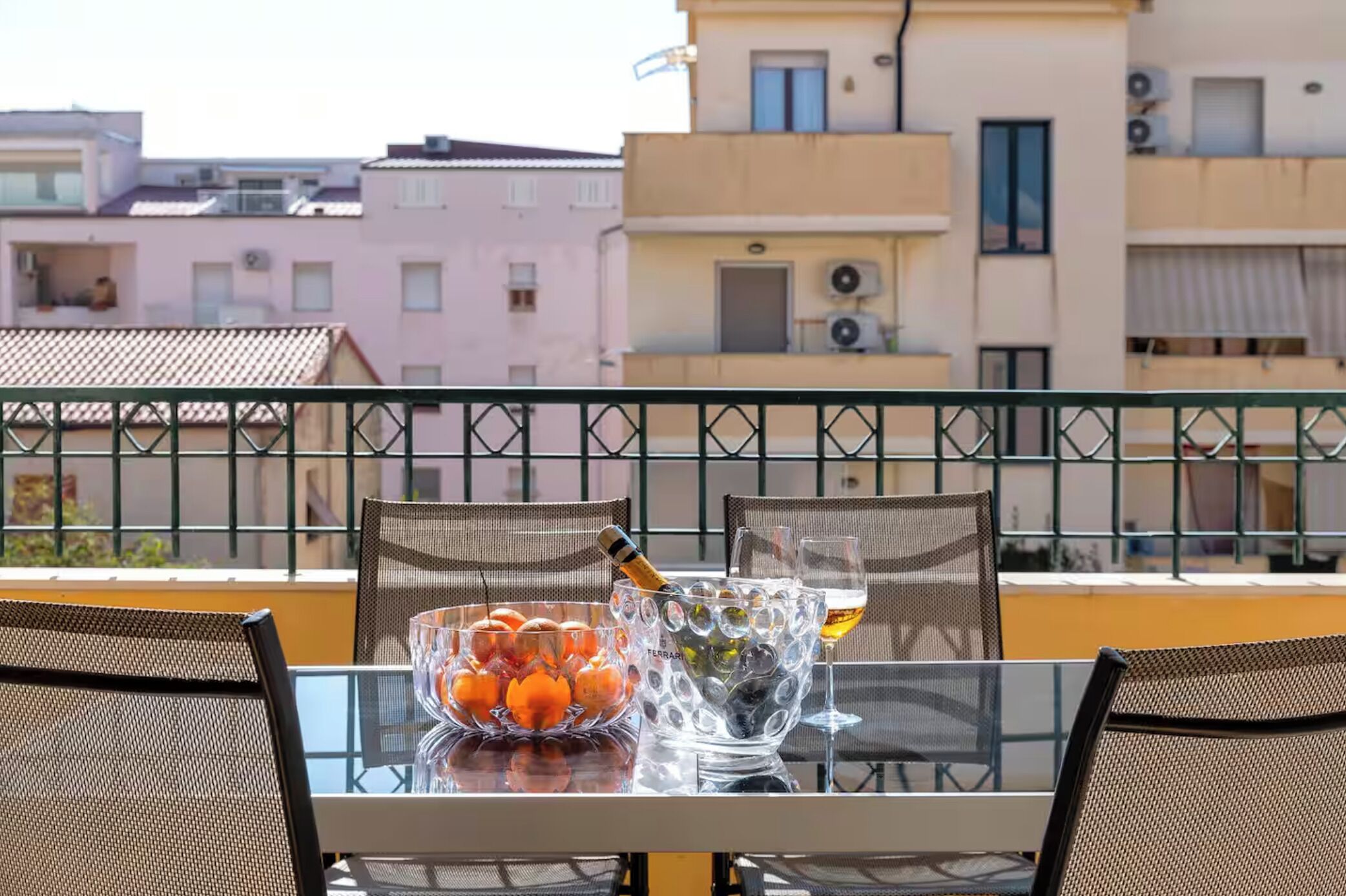 A balcony with a table set for two, featuring a bowl of oranges, a bottle of champagne, and a glass of wine.