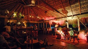 An indoor venue with a thatched roof, wooden floors, and string lights. Tables and chairs are arranged for guests. - Puerto Dreams H (Puerto Escondido)
