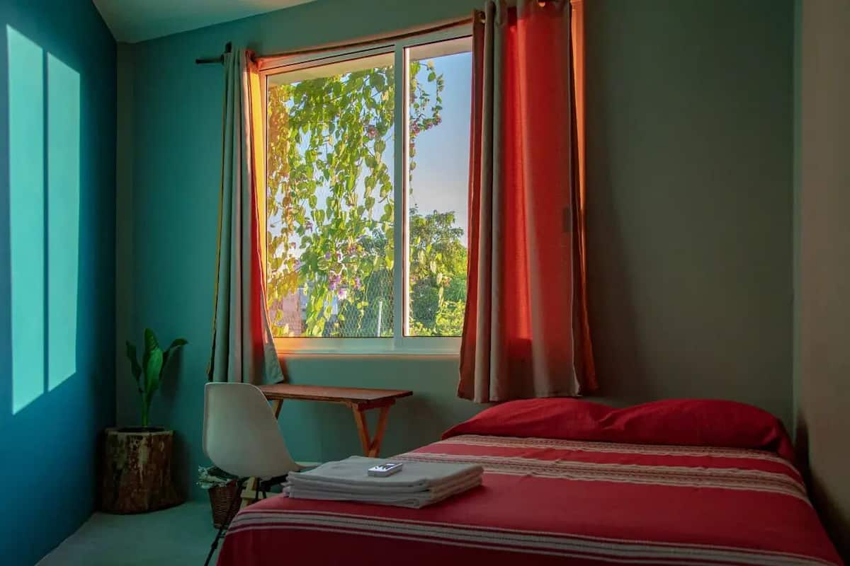 A bedroom with a bed, a desk, a chair, a window with curtains, and a plant.