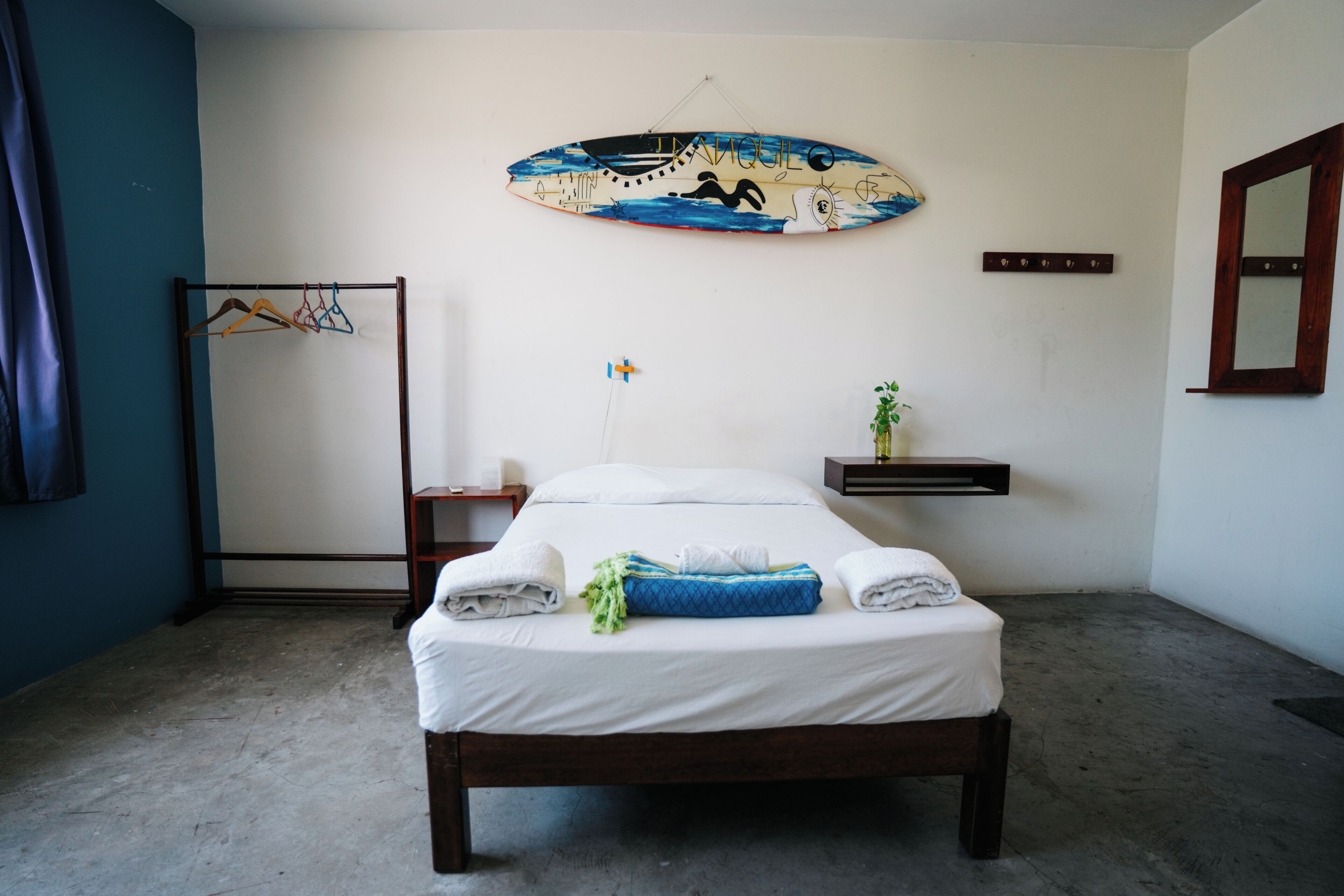 Een slaapkamer met een bed, een aan de muur bevestigd surfplank, een kledingroede en een klein plantje, plus een nachtkastje.
