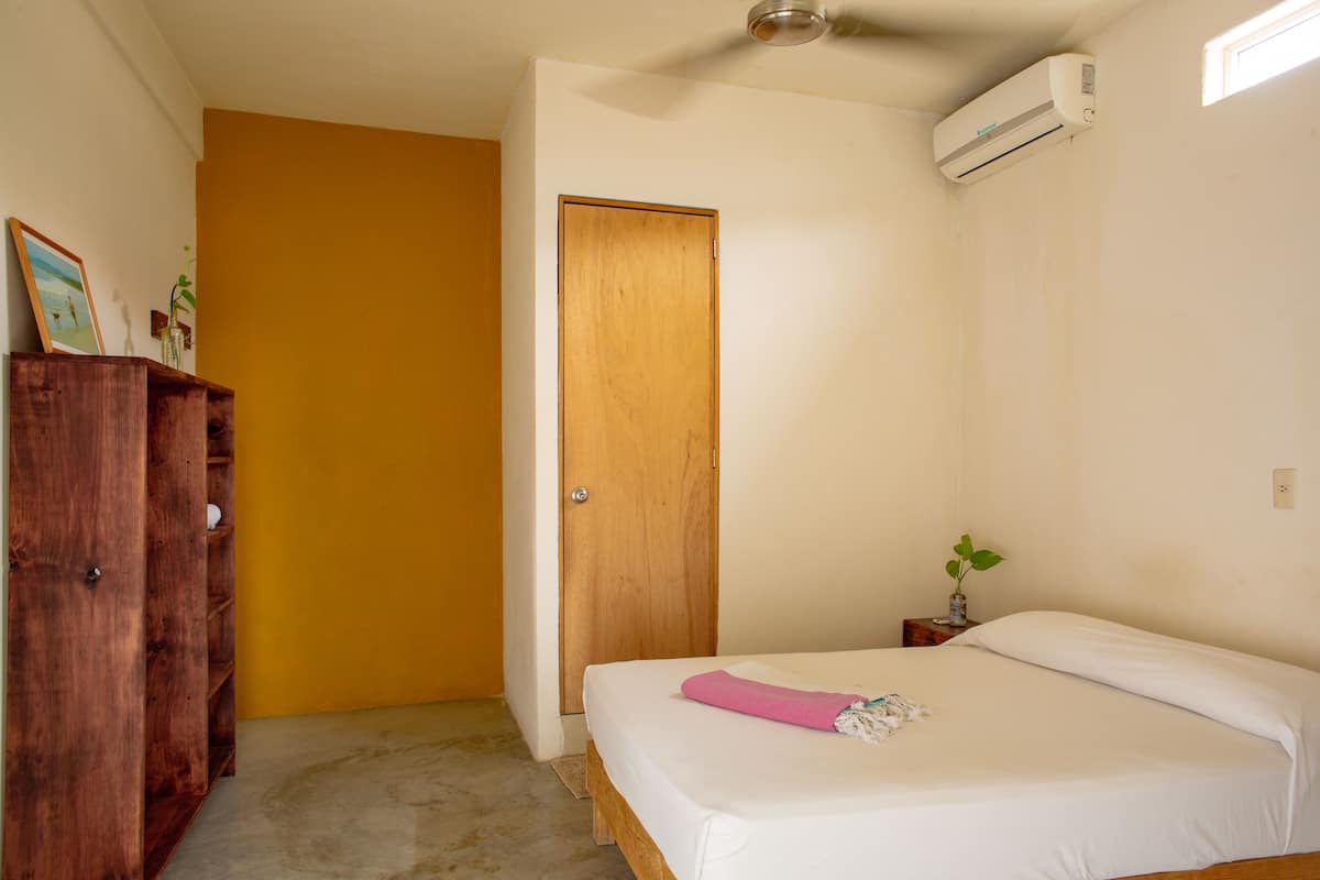 A bedroom with a bed, a wooden wardrobe, a door, and an air conditioning unit.