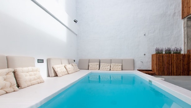 Un área de piscina interior moderna con interior blanco, un banco largo con cojines y una valla de madera con plantas.