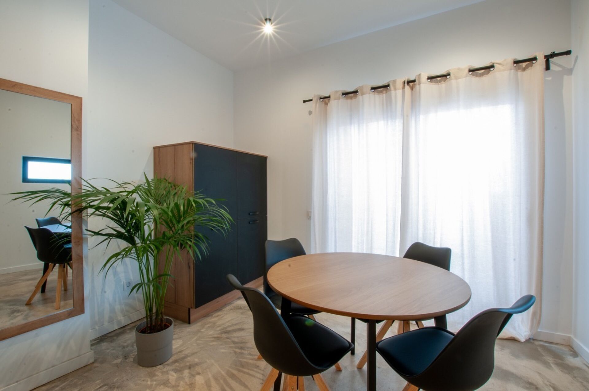 Une pièce moderne avec une table ronde en bois, quatre chaises noires, une plante en pot, une armoire et un grand miroir.