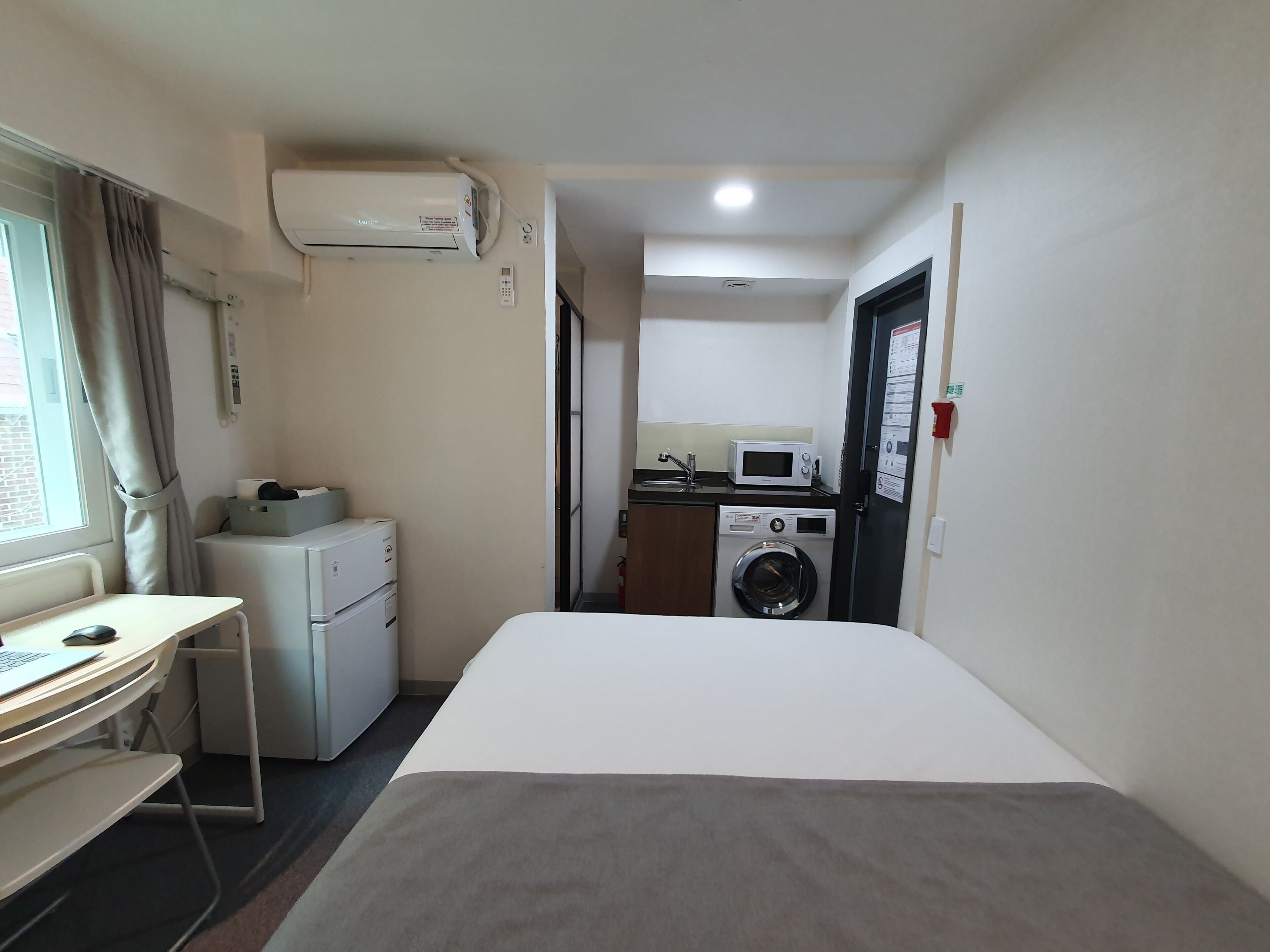 A hotel room with a bed, a desk with a computer, a washing machine, a microwave, and an air conditioner.