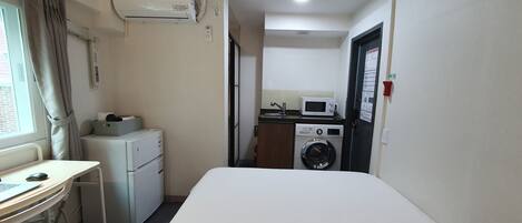 A hotel room with a bed, a desk with a computer, a washing machine, a microwave, and an air conditioner.