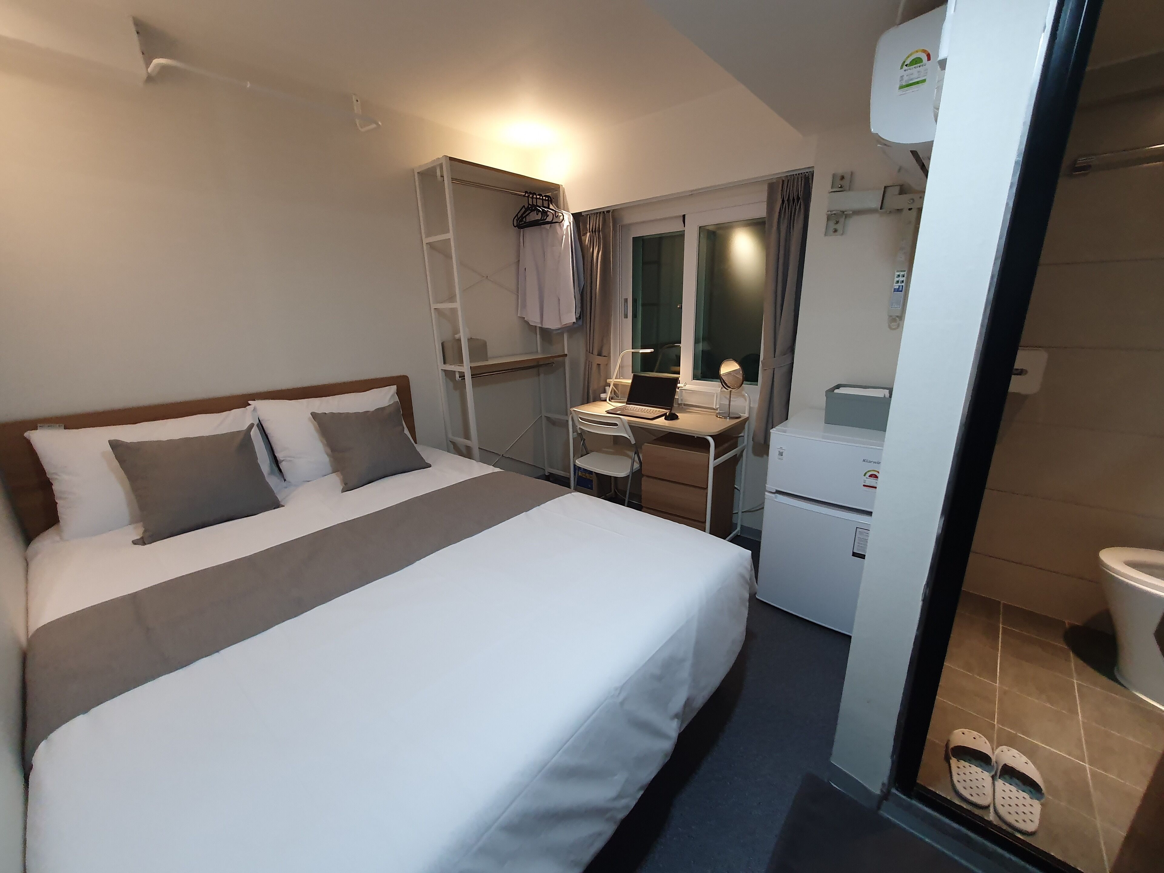 Habitación de hotel con cama, escritorio con computadora portátil, espejo, armario y una pequeña kitchenette.