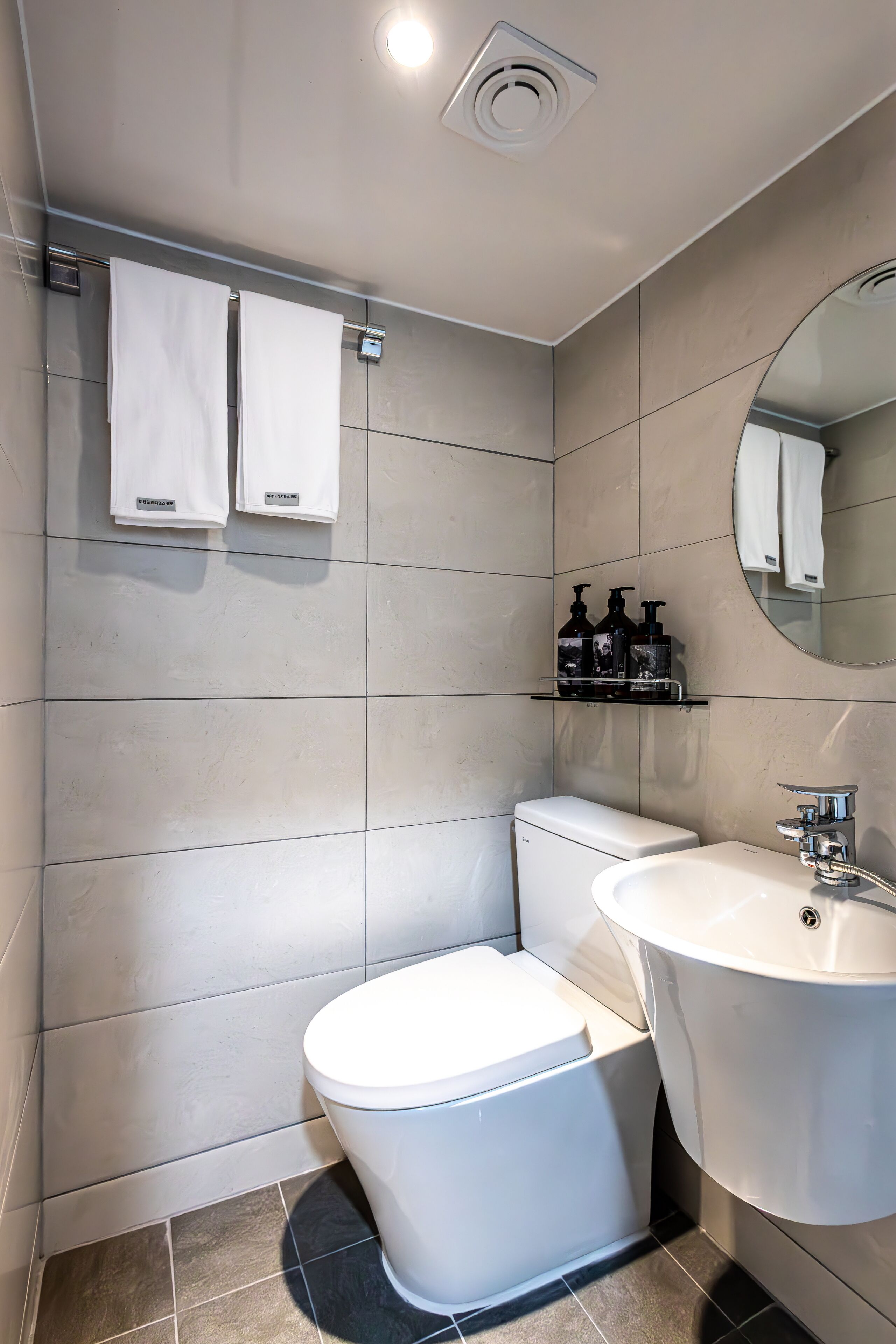 Ein modernes Badezimmer mit Toilette, Waschbecken und Spiegel. Zwei Handtücher hängen an einem Wäscheständer, und ein Regal enthält Pflegeprodukte.