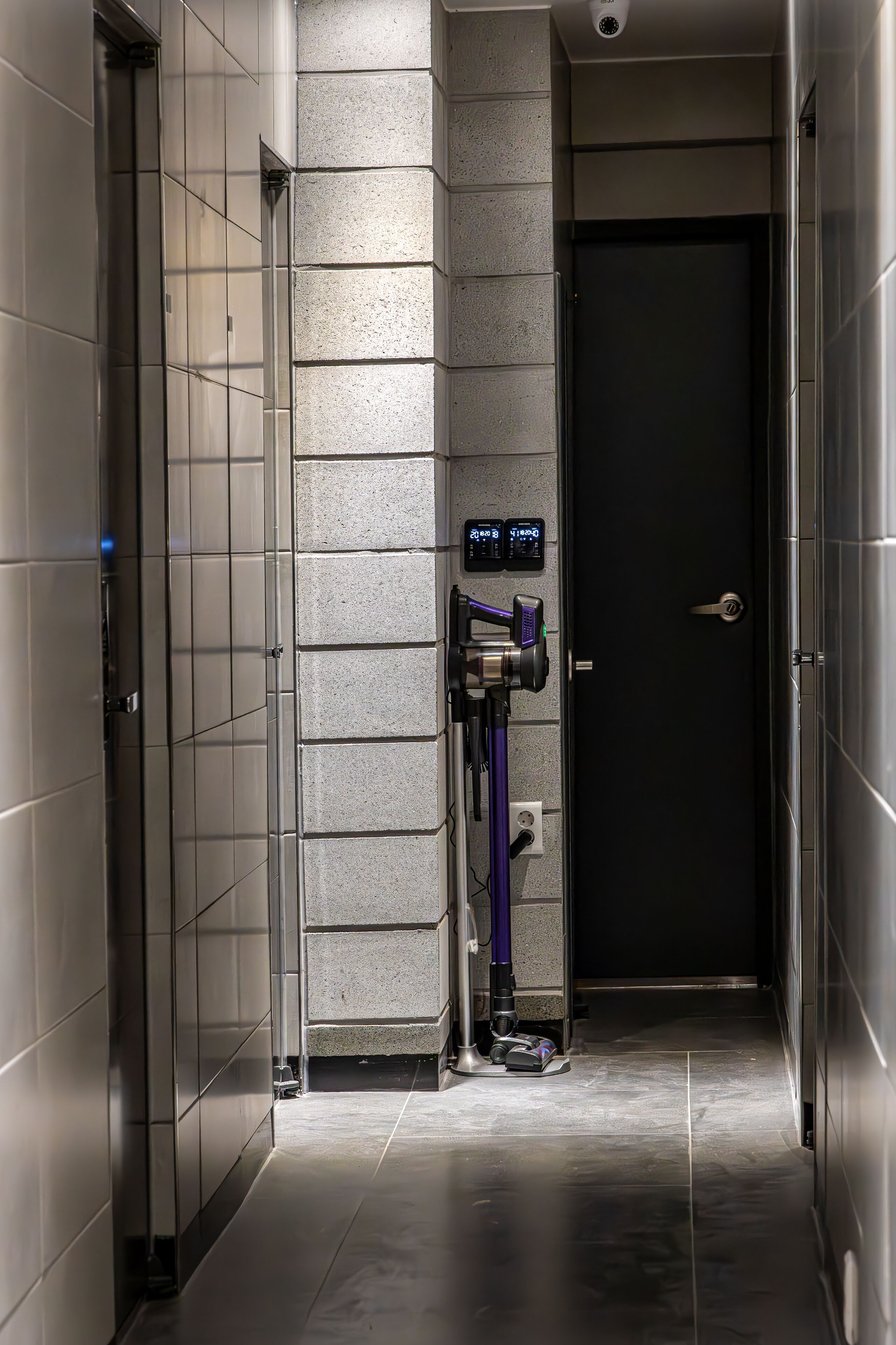 A narrow hallway with a door at the end, a security camera above, and a vacuum cleaner in the center.