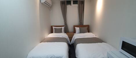 A hotel room with two single beds, a small desk with a TV, an air conditioning unit, and a window with curtains.