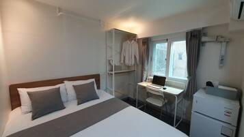 A hotel room with a bed, a desk with a laptop, a mini-fridge, and a window with curtains.