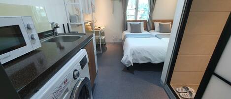 A compact hotel room with a kitchenette, a bed with two pillows, a small desk, and a window with curtains.