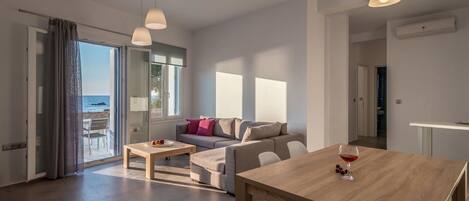 A modern living room with a sofa, a dining table, and a view of the sea through a glass door.