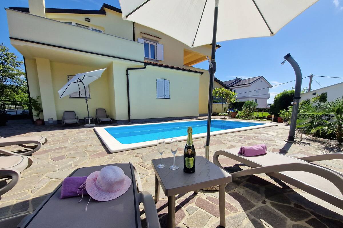 A poolside area with lounge chairs, a table with a bottle and glasses, and a large umbrella.