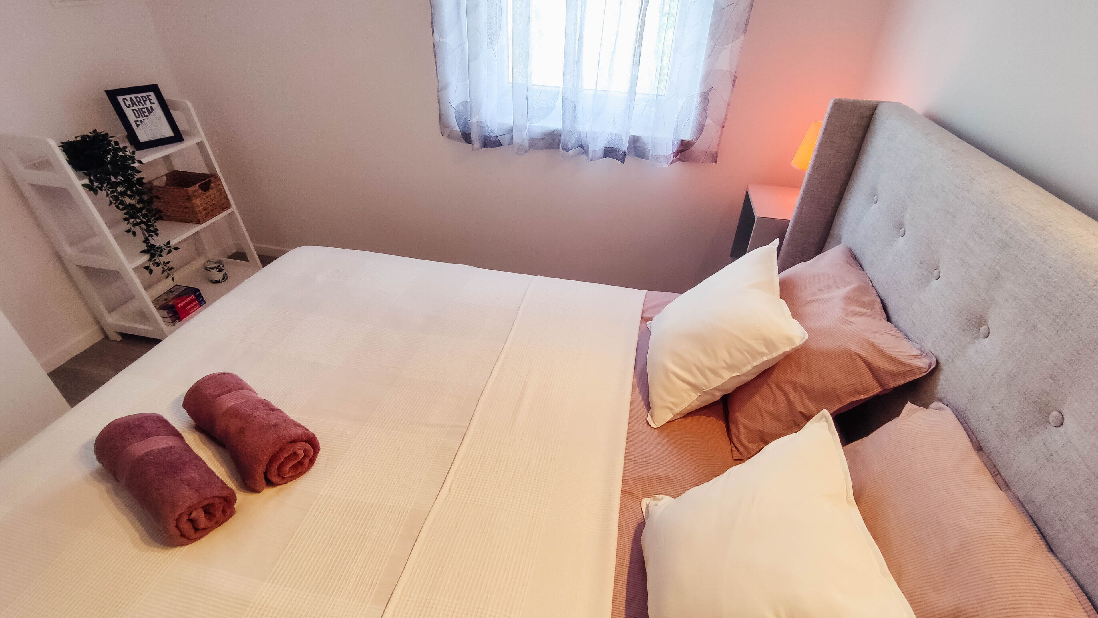 A neatly made bed with rolled towels, a bedside table with a lamp, and a window with sheer curtains.