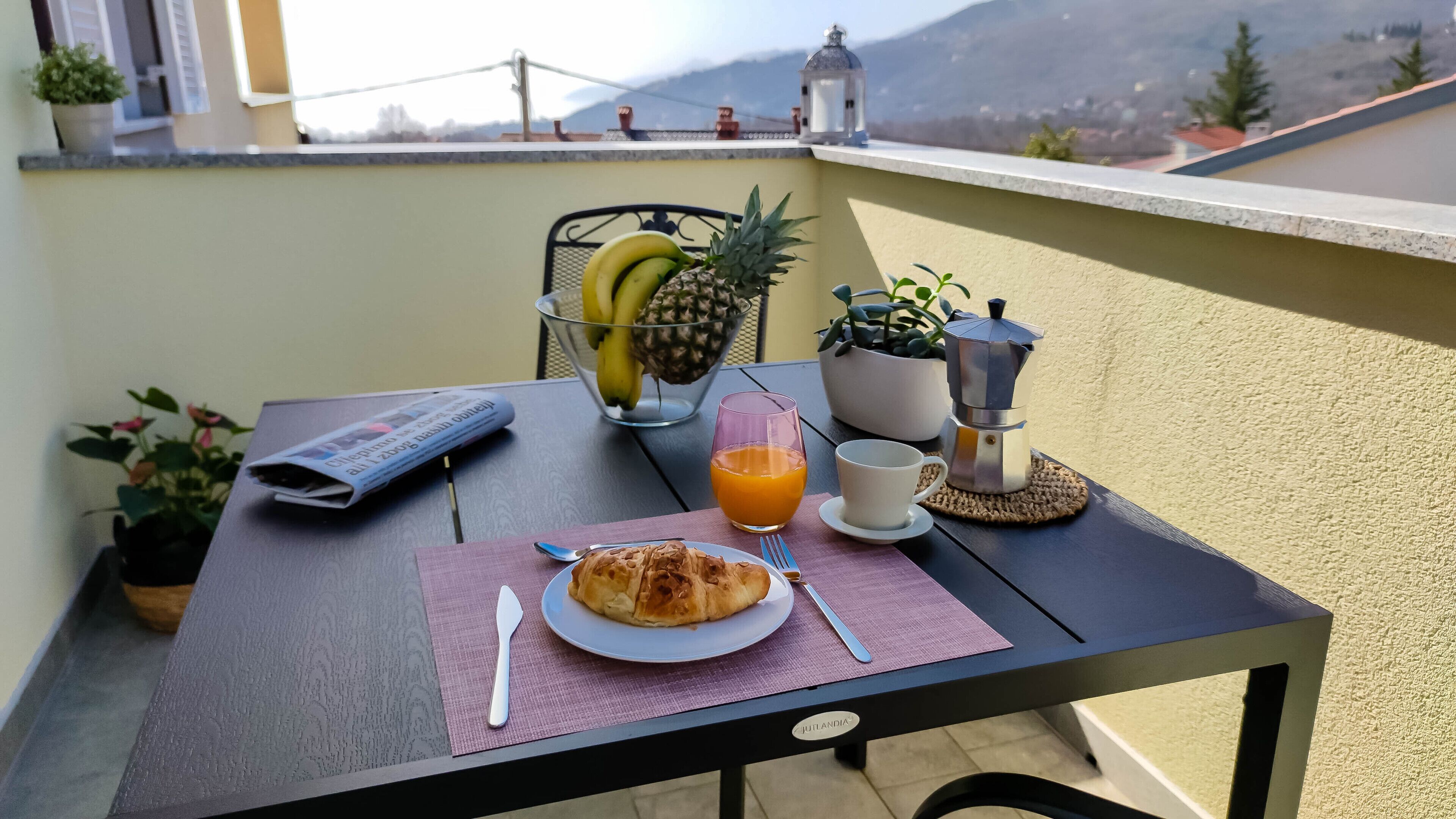 A table with a newspaper, a plate with food, a glass of orange juice, and a coffee maker on a balcony with a view of mountains.