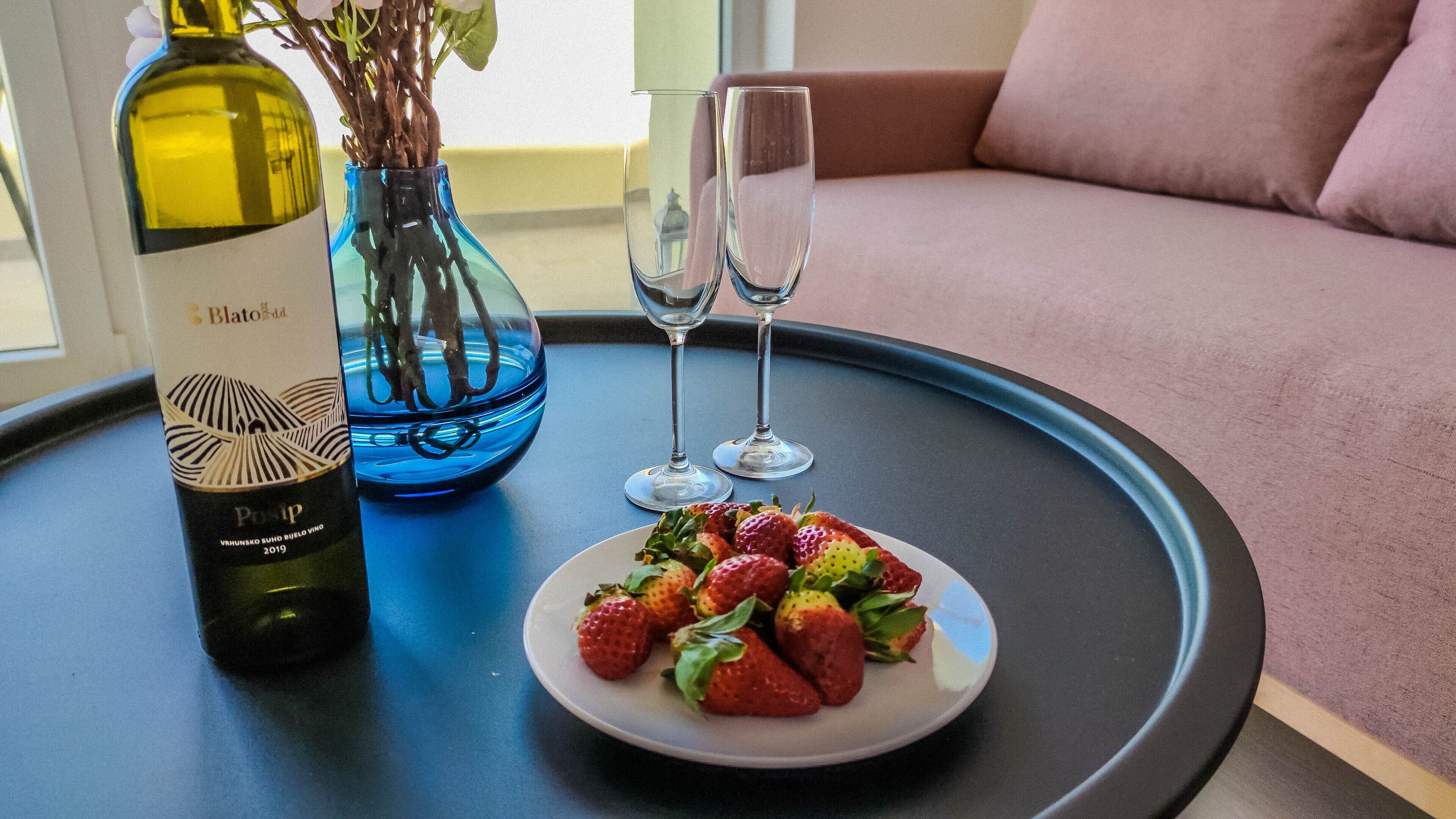 A table with a bottle of wine, a vase with branches, two glasses, and a plate of strawberries.